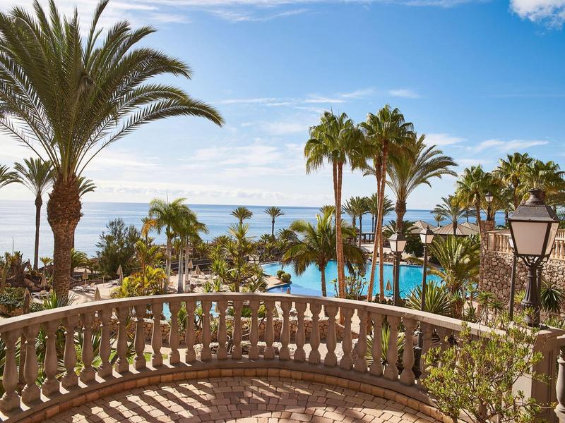 Vue depuis une terrasse avec balustrades sur une piscine, des palmiers et la mer par une journée ensoleillée.