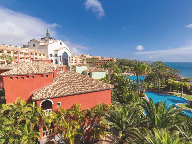 Complexe hôtelier avec bâtiments rouges, piscine et plantes tropicales sous ciel bleu au bord de la mer.