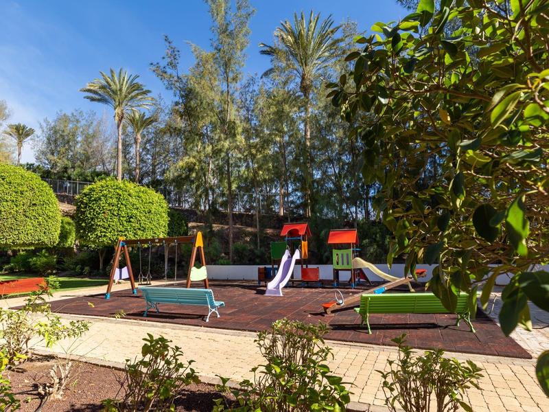 Aire de jeux pour enfants avec toboggans et balançoires dans un jardin d'hôtel ensoleillé et verdoyant.