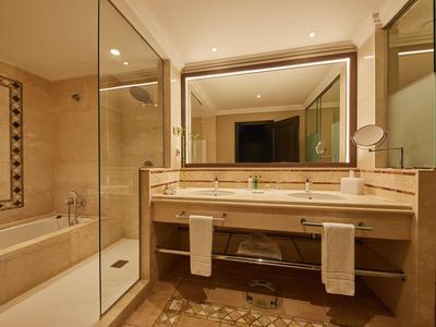 Hotelbadkamer met grote spiegel, dubbele wastafel, ligbad, douche en beige marmeren inrichting.