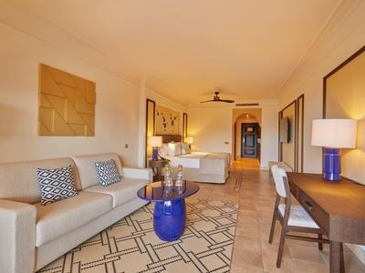 Moderne hotelsuite met bed, bank, bureau en blauwe accenten in warm licht.