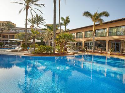 Großer blauer Pool mit Palmen und zweistöckigem Hotelgebäude bei klarem Himmel.