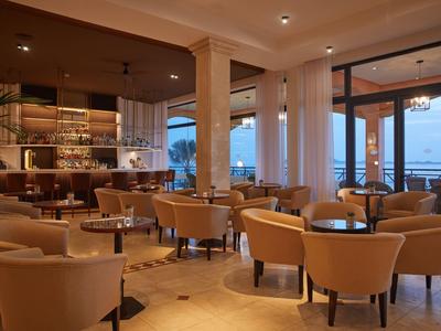 Gezellige hotelbar met beige stoelen, houten tafels en uitzicht op zee door grote ramen.