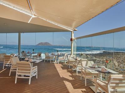 Buitenterras met witte stoelen en tafels met uitzicht op strand en zee onder een luifel.