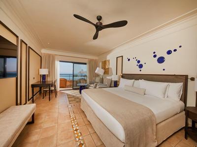 Ruime hotelkamer met groot bed, plafondventilator, bureau en balkon met zeezicht.