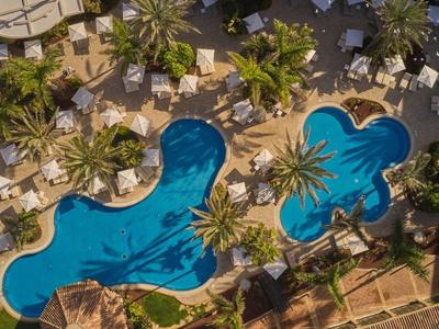 Luchtfoto van een resortzwembad met twee uniek gevormde zwembaden omgeven door ligstoelen en palmbomen.