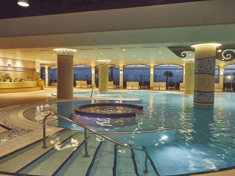 Overdekt hotelzwembad met ronde jacuzzi en panoramische ramen met uitzicht op zee.