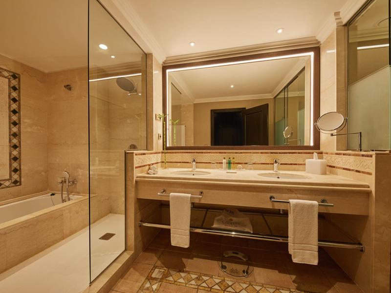 Hotelbadkamer met grote spiegel, dubbele wastafel, ligbad, douche en beige marmeren inrichting.