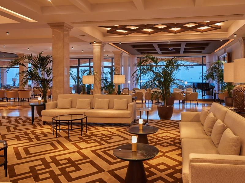 Ruime hotel lobby met beige banken, patroon tapijt, palmbomen en grote ramen.