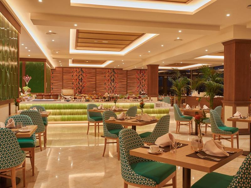 Moderne restaurantinterieur met groene stoelen, houten tafels en warme LED-plafondverlichting.