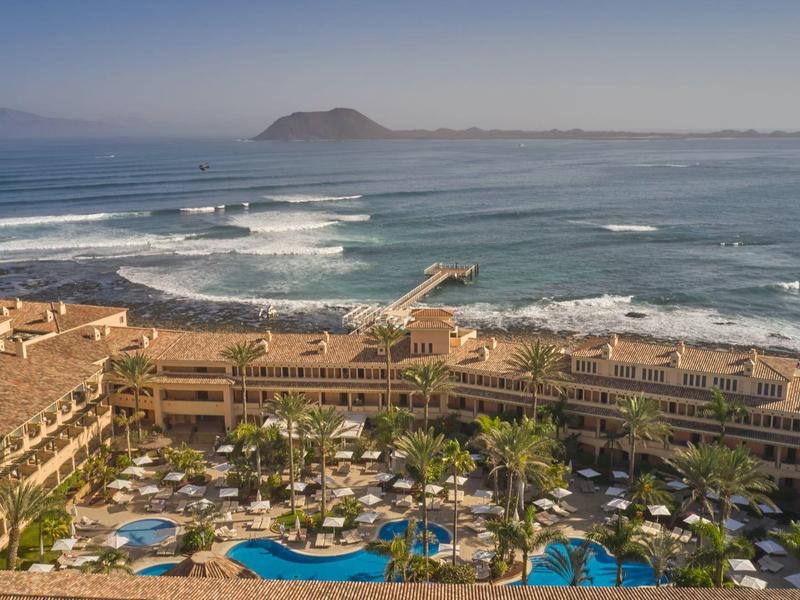 Groot resort aan de oceaan met meerdere zwembaden, palmbomen en een pier die de zee in loopt.