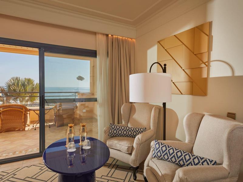 Gezellige hotelkamer met fauteuils en balkon met uitzicht op de oceaan bij zonsondergang.