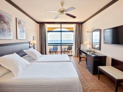 Jasny pokój hotelowy z podwójnymi łóżkami, balkonem i widokiem na morze.