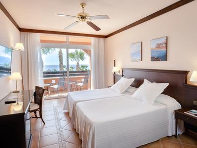 Jasny pokój hotelowy z dwoma łóżkami, balkonem i widokiem na palmy.