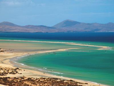 Plaża z turkusową wodą i wydmami, góry na horyzoncie pod niebieskim niebem.