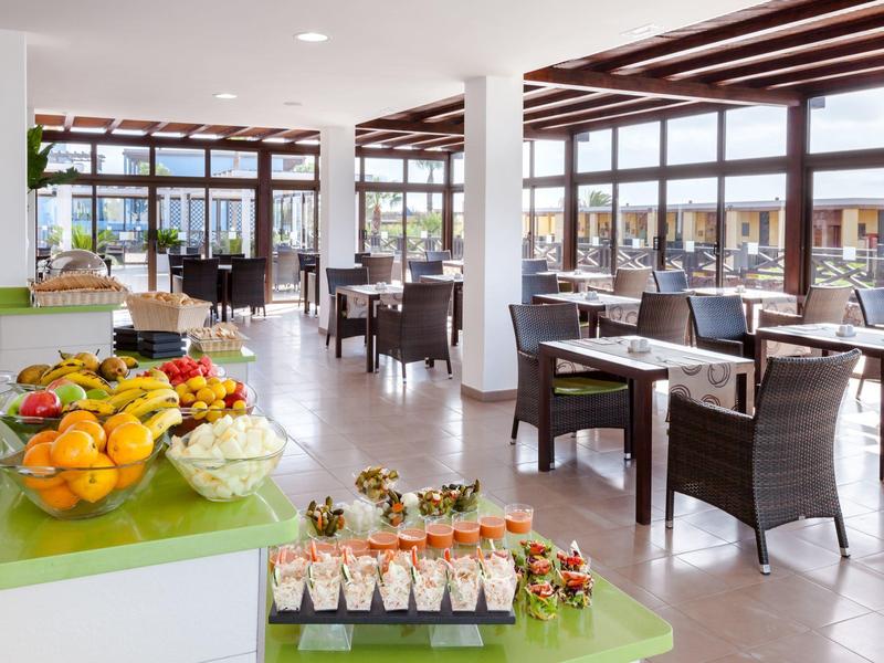 Jasna sala restauracyjna z bufetem owoców i przekąsek, duże okna.