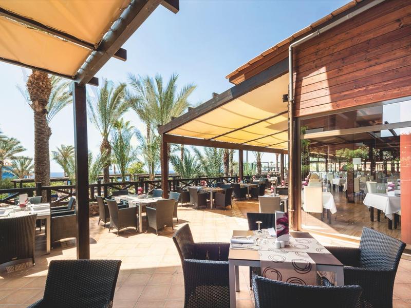 Taras restauracyjny z stołami, krzesłami pod drewnianymi zadaszeniami i palmami w oddali.