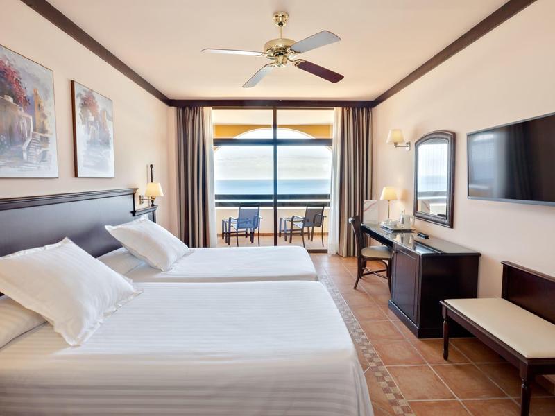 Jasny pokój hotelowy z podwójnymi łóżkami, balkonem i widokiem na morze.