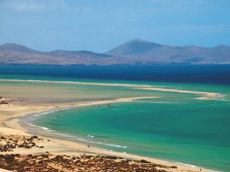 Plaża z turkusową wodą i wydmami, góry na horyzoncie pod niebieskim niebem.