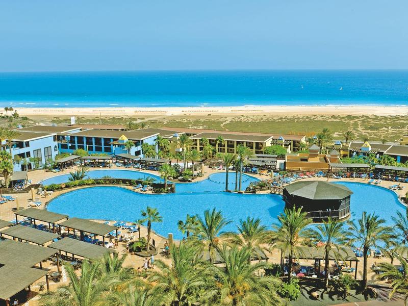 Widok na nadmorski resort z basenem, palmami i plażą przy turkusowym morzu.