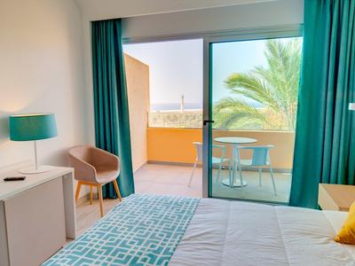 Camera d'hotel luminosa con letto, sedia, lampada da tavolo e balcone con vista sul mare.