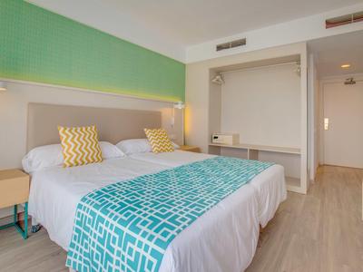 Camera d'hotel moderna con due letti, mobili in legno chiaro e accenti turchesi.