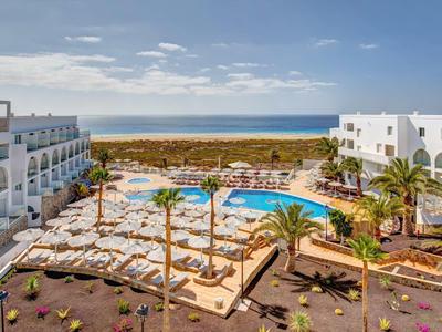 Hotel con due piscine, palme e vista mare in una giornata soleggiata.