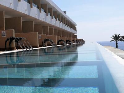 Piscina a sfioro moderna con cascate davanti a un edificio alberghiero con vista sul mare e palme.