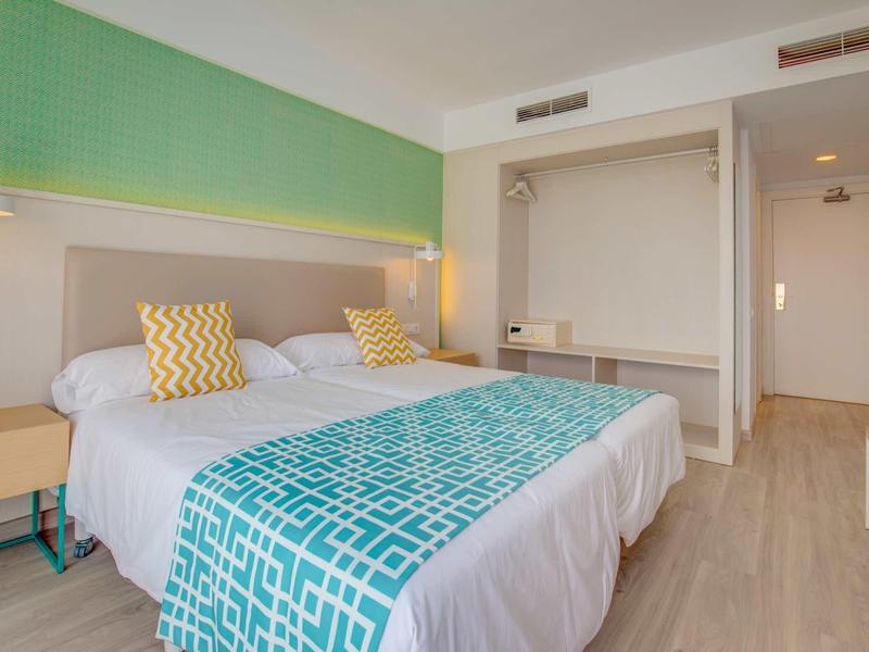 Camera d'hotel moderna con due letti, mobili in legno chiaro e accenti turchesi.