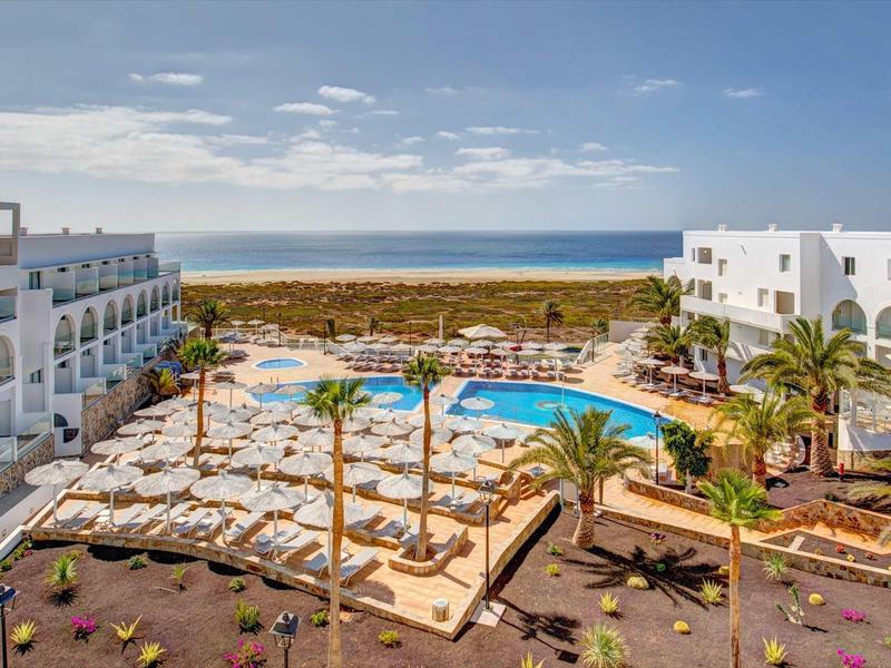 Hotel con due piscine, palme e vista mare in una giornata soleggiata.
