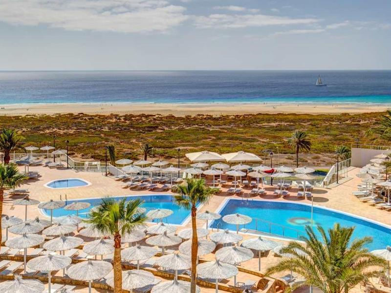 Piscina dell'hotel con ombrelloni, palme e vista sul mare sotto un cielo nuvoloso.