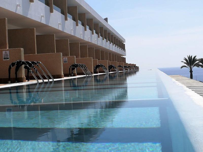 Piscina a sfioro moderna con cascate davanti a un edificio alberghiero con vista sul mare e palme.
