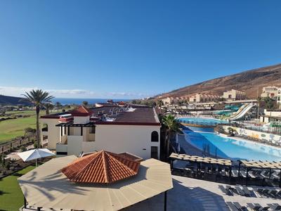 Widok na hotel z dużym basenem, parasolami przeciwsłonecznymi i tłem wzgórza pod czystym niebem.