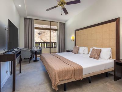 Pokój hotelowy z podwójnym łóżkiem, telewizorem, krzesłem, balkonem i wentylatorem sufitowym.