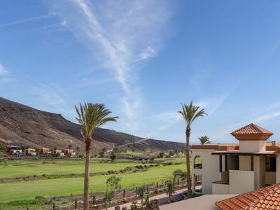 Vue d'un complexe avec piscine, palmiers et montagnes sous un ciel bleu.
