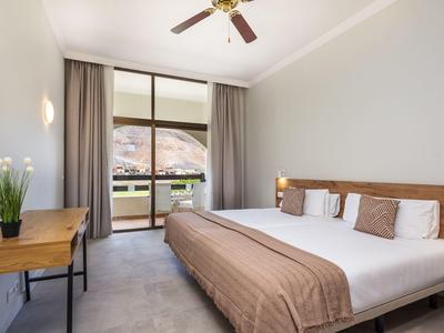 Jasny pokój hotelowy z dużym łóżkiem, drewnianym biurkiem i widokiem na balkon i góry.