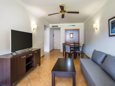 Prosty salon z szarą sofą, telewizorem, stołem kawowym oraz jadalnią w tle.