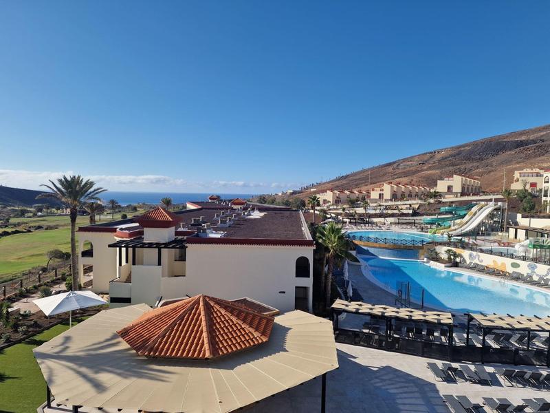 Widok na hotel z dużym basenem, parasolami przeciwsłonecznymi i tłem wzgórza pod czystym niebem.