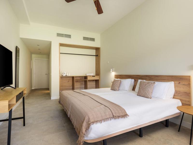 Jasny pokój hotelowy z łóżkiem podwójnym, telewizorem na ścianie i jasnym drewnianym meblem.