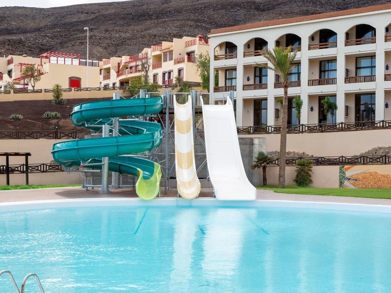 Niebieski basen z zjeżdżalniami i hotelem w tle pod jasnym niebem.