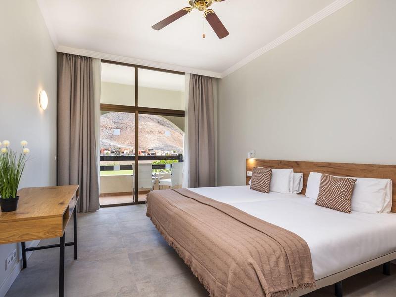 Jasny pokój hotelowy z dużym łóżkiem, drewnianym biurkiem i widokiem na balkon i góry.