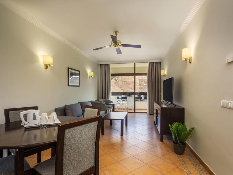 Nowoczesny hotelowy salon z kanapą, telewizorem i stołem przy drzwiach balkonowych.