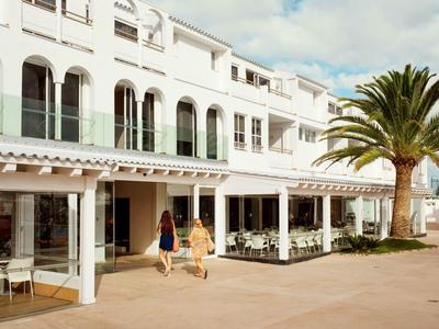 Edificio de hotel blanco con grandes ventanas, terraza y palmeras en un día soleado.