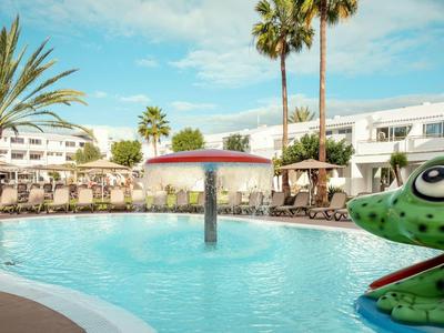 Piscina del hotel con tumbonas, sombrillas y palmeras bajo el cielo azul.