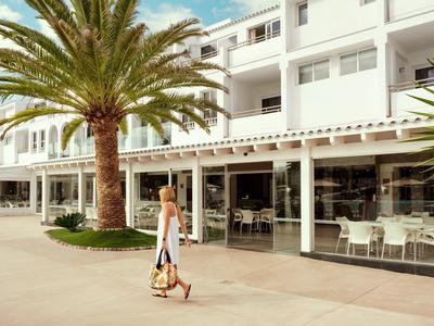 Mujer con bolso camina delante de un hotel moderno con palmeras y terraza exterior.