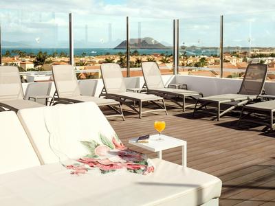 Terraza en la azotea con tumbonas blancas, mesa, bebida y vista a la ciudad y puerto.