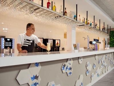 Bartender prepara bebidas en bar moderna de hotel con estantes de vidrio y barra de mármol.