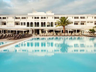 Grand bâtiment d'hôtel à façade blanche avec une grande piscine extérieure sous un ciel nuageux.