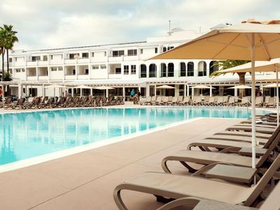 Piscine d'hôtel avec transats et parasols devant un bâtiment blanc à plusieurs étages sous un ciel nuageux.