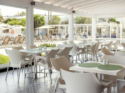 Café moderno y luminoso con mesas y sillas blancas, vista al área exterior con tumbonas.